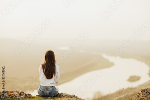 Back view of woman sitting on top of the hill and looking at river in evening sunset. Young traveler enjoying time in nature. Lifestyle travel, relaxation and freedom concept. Copy space. 