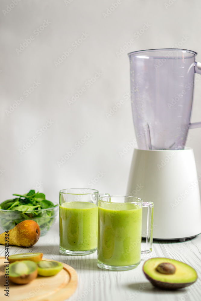 Green smoothie in glass with pear, spinach, kiwi, avocado and blender on white wooden table. Copy space. Green smoothie in glass with pear, spinach, kiwi, avocado and blender on white wooden table. Copy space.