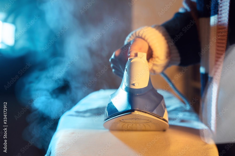 Fototapeta premium Close up on female caucasian hand woman holding steam iron ironing cloth dress in dark room at home in night or evening