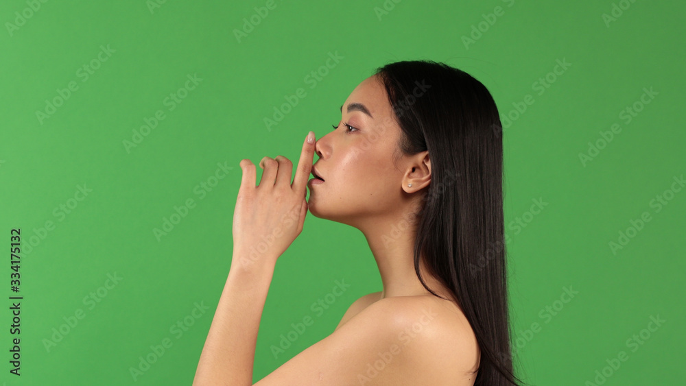 Portrait of asian girl standing in profile holding index finger near her mouth. The girl gestures to be quiet and no one talks Portrait of asian girl standing in profile holding index finger near her mouth. The girl gestures to be quiet and no one talks