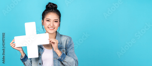 Banner of Happy asian woman smiling and holding plus or add sign on copy space blue background. Cute asia girl smiling wearing casual jeans shirt and showing join sign for increse and more concept photo