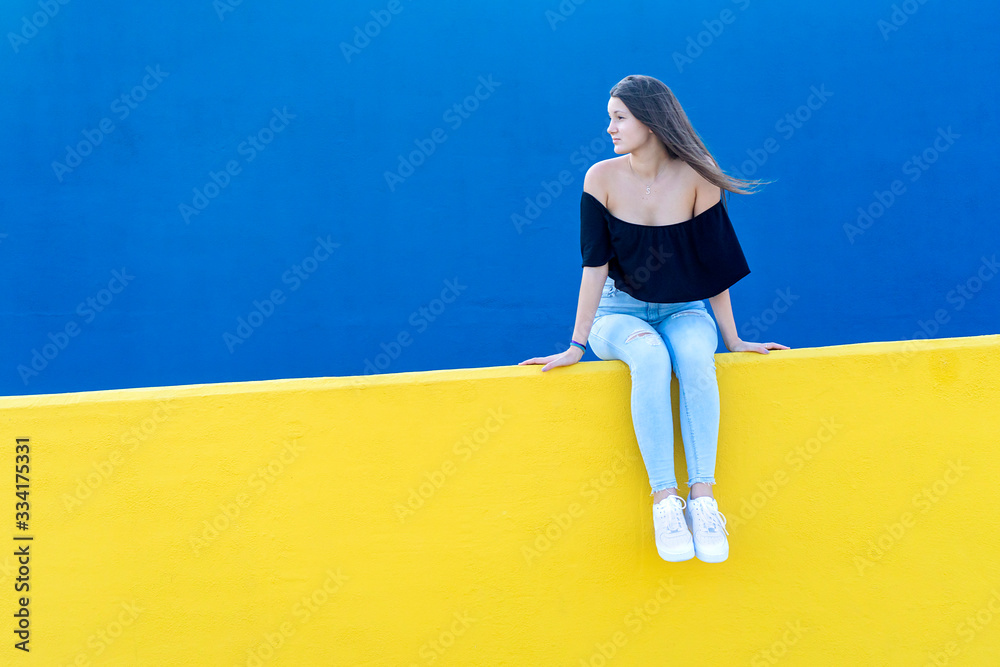 Young beautiful woman with long hair sitting on yellow wall while looking pensive aside in a sunny day Young beautiful woman with long hair sitting on yellow wall while looking pensive aside in a sunny day