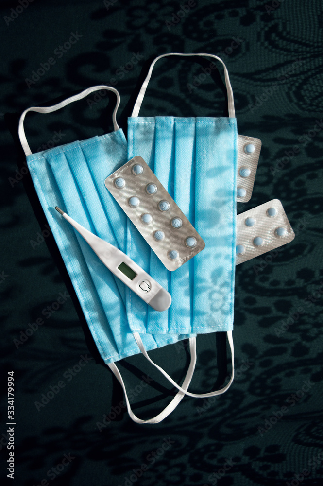 Vertical photo of two blue medical face masks, thermometer and tablets on the dark emerald background with shadows ornaments againts virus covid-2019. Heath care concept, stop coronavirus