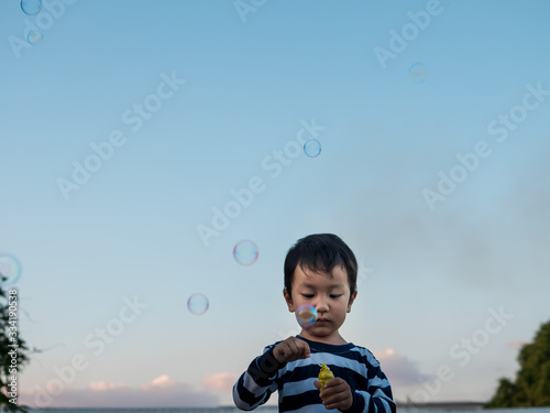 Asian child boy playing bubbles outdoor with happy face with nature blue sky background, bubbles in the air. Young kid have fun in relaxing holiday, freedom concept.