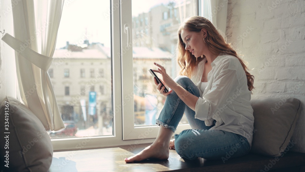 Charming young woman sitting on a windowsill at home and texting on her phone communication female looking message cellphone cheerful smile use internet modern smartphone - obrazy, fototapety, plakaty 