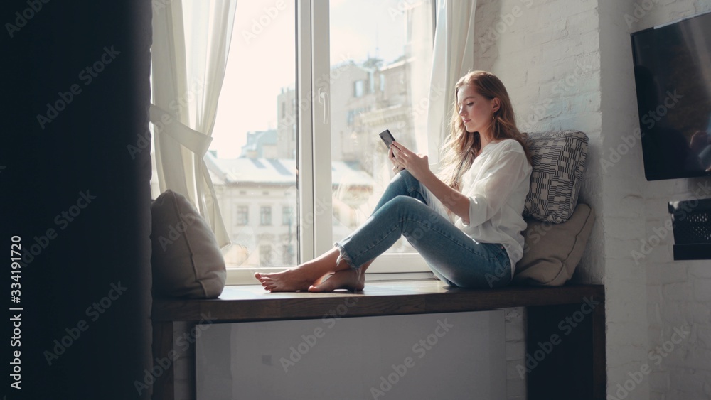 Cheerful young woman sitting on a windowsill at home and texting on her phone communication female looking message cellphone cheerful smile use internet modern smartphone portrait - obrazy, fototapety, plakaty 