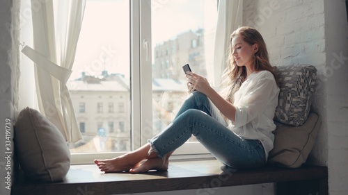 Pretty young woman sitting on a windowsill at home and texting on her phone communication female looking message cellphone cheerful smile use internet modern smartphone portrait photo
