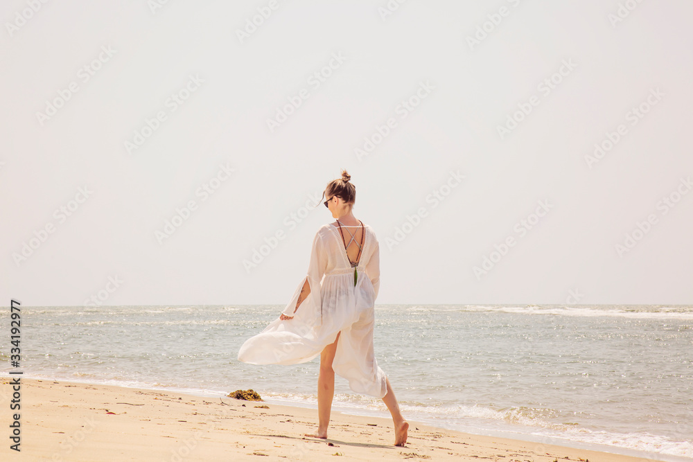 Woman in white dress and mala necklace walking away on the sunny beach by the sea Woman in white dress and mala necklace walking away on the sunny beach by the sea