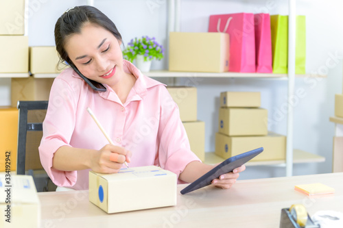 Young beautiful happy asian business woman owner of SME online using laptop receive order from customer with parcel box packaging at her startup home office, online business seller and delivery