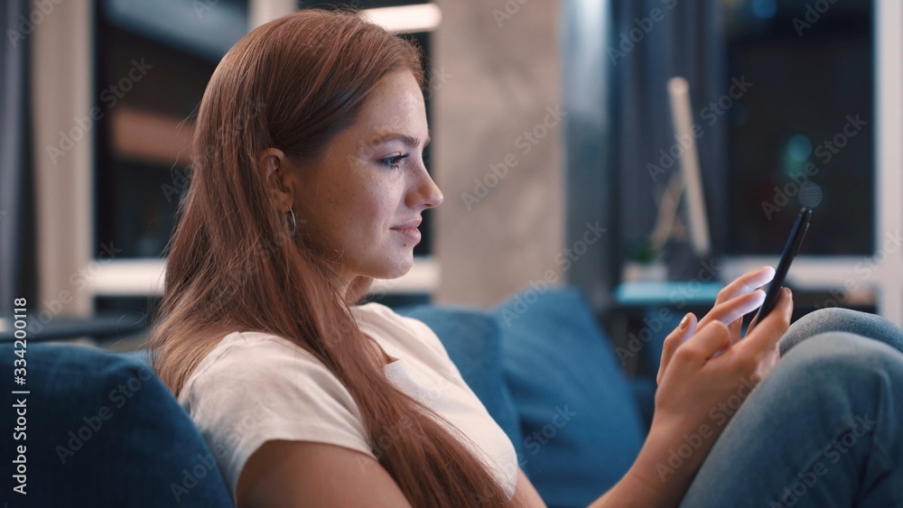 Relaxing cheerful young woman with golden hair sitting on blue sofa uses phone smile at modern apartment texting share messages applications on social media enjoy feel happy mobile technology evening