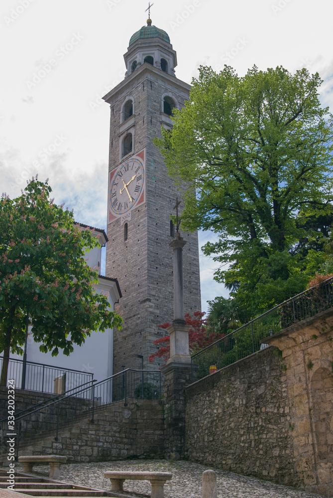 Church yard with green trees and stone benches, tall bell tower with cross at the top and clock. Sunny summer day in Lugano, Switzerland. Church yard with green trees and stone benches, tall bell tower with cross at the top and clock. Sunny summer day in Lugano, Switzerland.