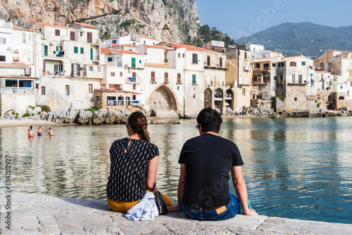 Couple sits on the quay wall and enjoys the view of the coastal town of Cefalu; Italy
