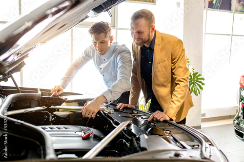affable handsome caucasian consultant and client in dealership. young business man came to buy new automobile, happy future owner of representative car, examining car hood