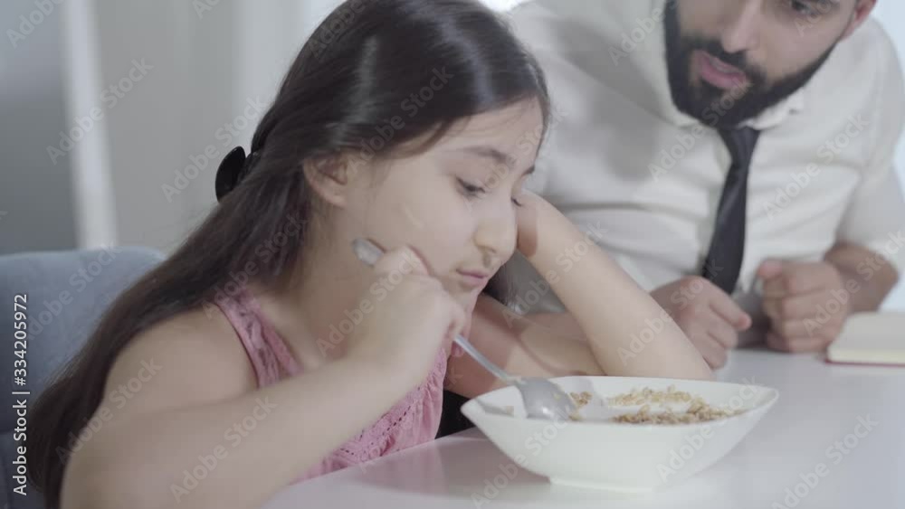Blurred young Middle Eastern man yelling at sad little girl mixing porridge with spoon. Portrait of hurrying father arguing with disobedient daughter in the morning. Lifestyle, breakfast, parenthood. Blurred young Middle Eastern man yelling at sad little girl mixing porridge with spoon. Portrait of hurrying father arguing with disobedient daughter in the morning. Lifestyle, breakfast, parenthood.