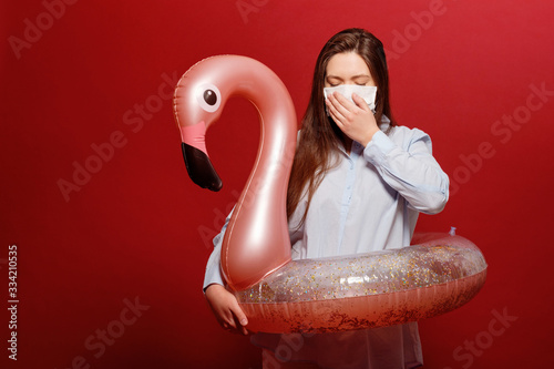 office employee young beautiful woman on red background in protective medical mask hugs swimming circle pink flamingo in depression, pandemic, vacation cancellation, sneeze © denisval