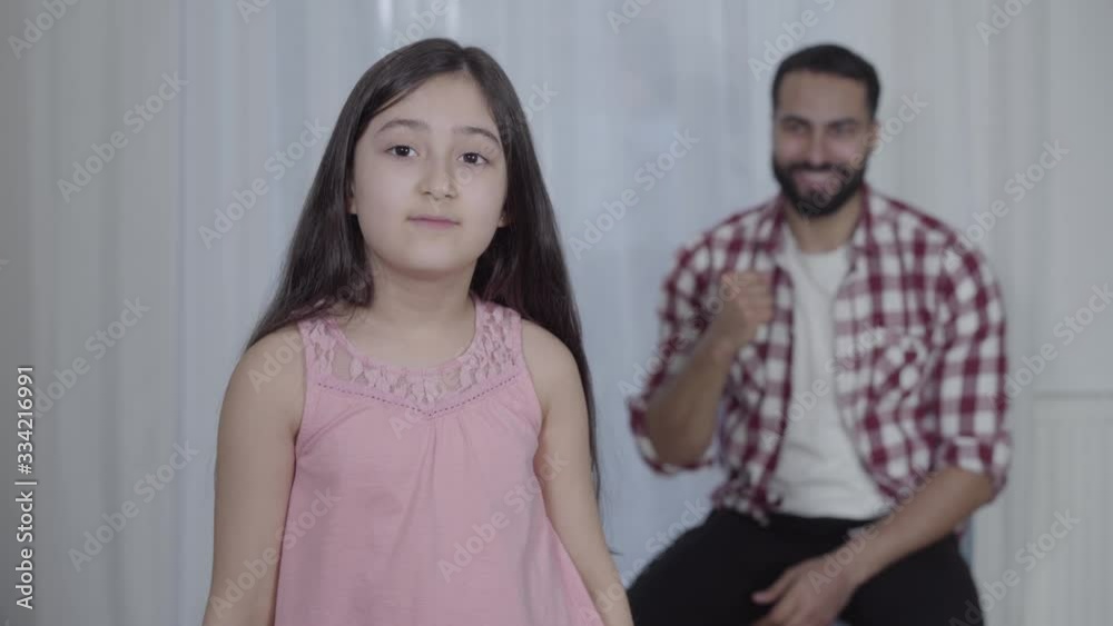 Confident Middle Eastern girl in boxing gloves kicking hands as proud man smiling at the background. Little sportive daughter posing with young handsome father indoors. Self-defense, sport, lifestyle. Confident Middle Eastern girl in boxing gloves kicking hands as proud man smiling at the background. Little sportive daughter posing with young handsome father indoors. Self-defense, sport, lifestyle.