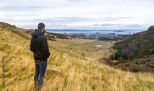 young man standing on mazing yellow and green mountain landscape watching a city at the horizon © Alex
