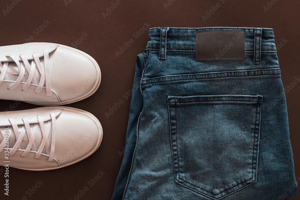 A set of white leather sneakers, belt and blue jeans on brown background. Copy space. Top view.