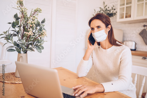 European sick woman in medical mask speaking smartphone and working on a laptop in kitchen home quarantine isolation Covid-19 pandemic Corona virus. Online work, stay home. Thermometer and pills