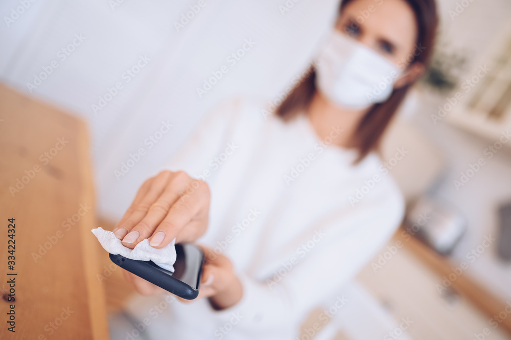 Fototapeta premium Unrecognizable woman in face protection mask cleaning mobile phone to eliminate germs Covid-19 by hand sanitizer, using cotton wool with alcohol to wipe to avoid contaminating with Corona virus. 