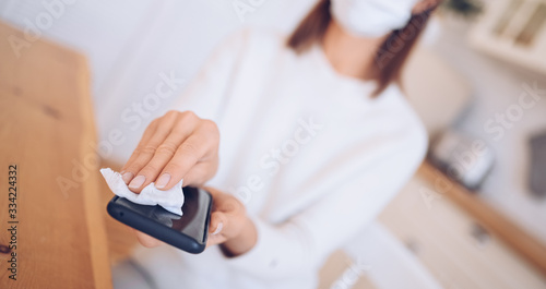 Unrecognizable woman in face protection mask cleaning mobile phone to eliminate germs Covid-19 by hand sanitizer  using cotton wool with alcohol to wipe to avoid contaminating with Corona virus. 