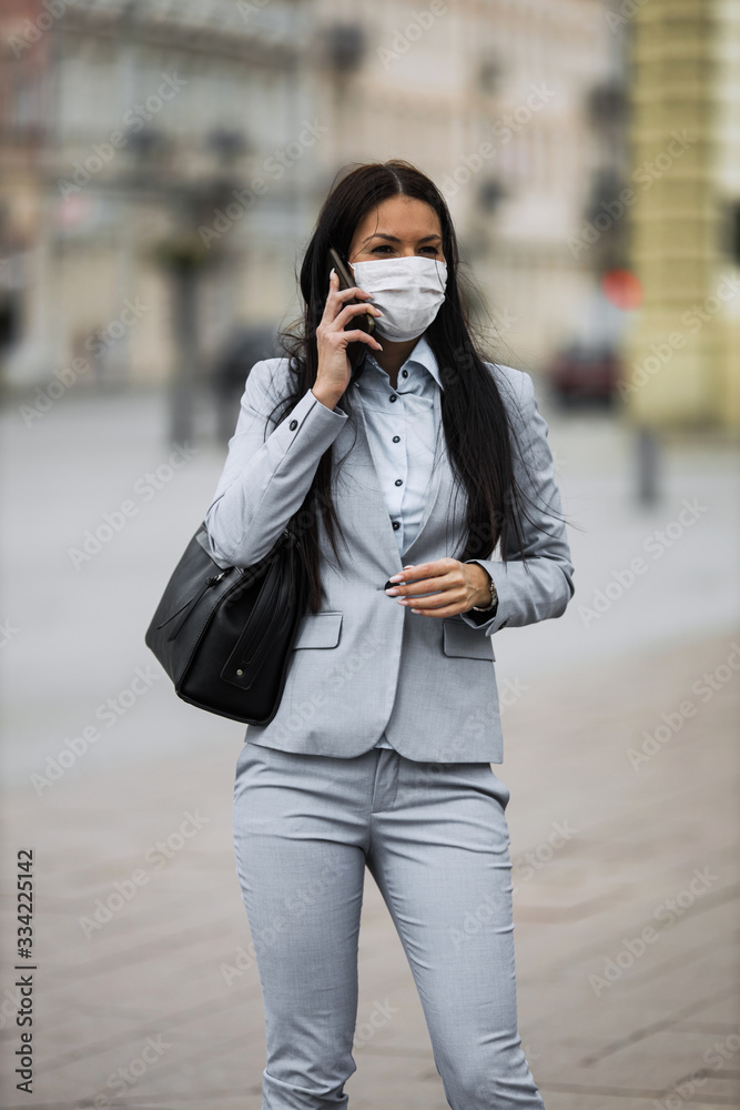 Young and elegant business woman on empty city street using smart phone and wearing protective mask to protect herself from dangerous flu or virus. Corona virus or Covid-19 concept. Young and elegant business woman on empty city street using smart phone and wearing protective mask to protect herself from dangerous flu or virus. Corona virus or Covid-19 concept.