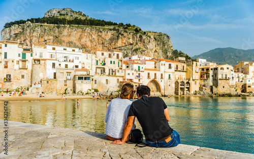 Couple sits on the quay wall and enjoys the view of the coastal town of Cefalu; Italy