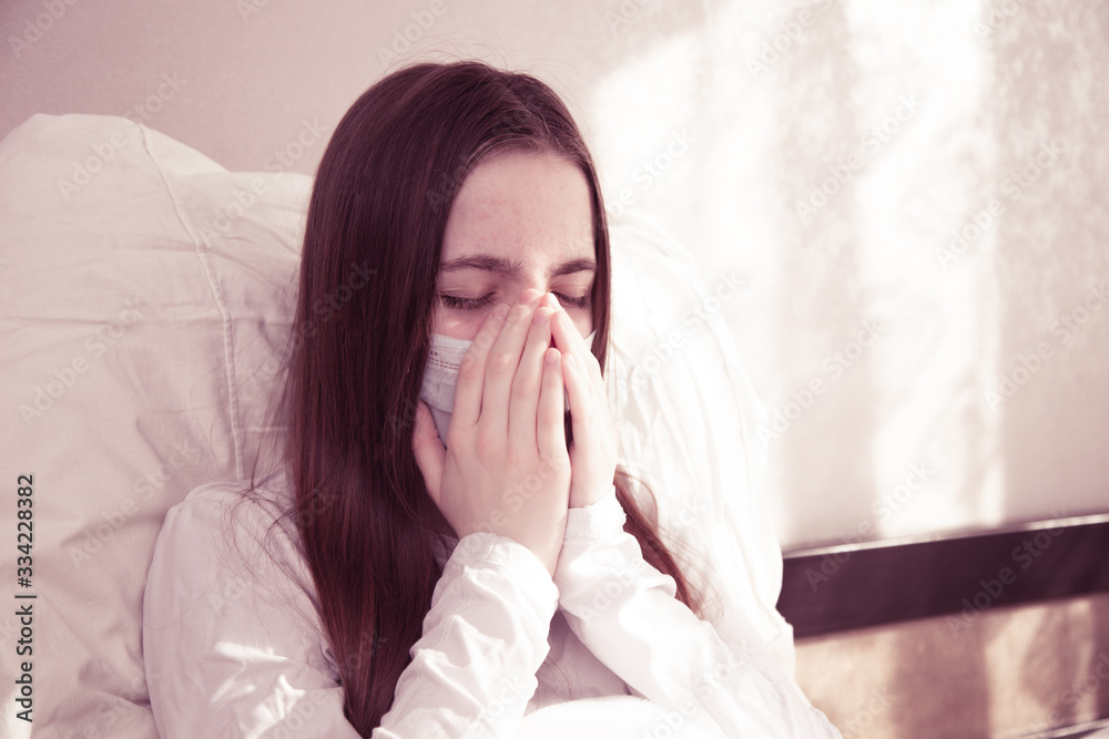 a young girl with brown hair is sitting at home in quarantine, a young girl is sitting in bed in a mask from the spread of the virus, infection and coughing in the palm of her hands. stay at home a young girl with brown hair is sitting at home in quarantine, a young girl is sitting in bed in a mask from the spread of the virus, infection and coughing in the palm of her hands. stay at home