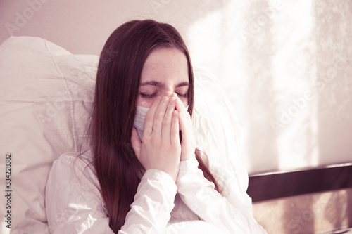 a young girl with brown hair is sitting at home in quarantine, a young girl is sitting in bed in a mask from the spread of the virus, infection and coughing in the palm of her hands. stay at home