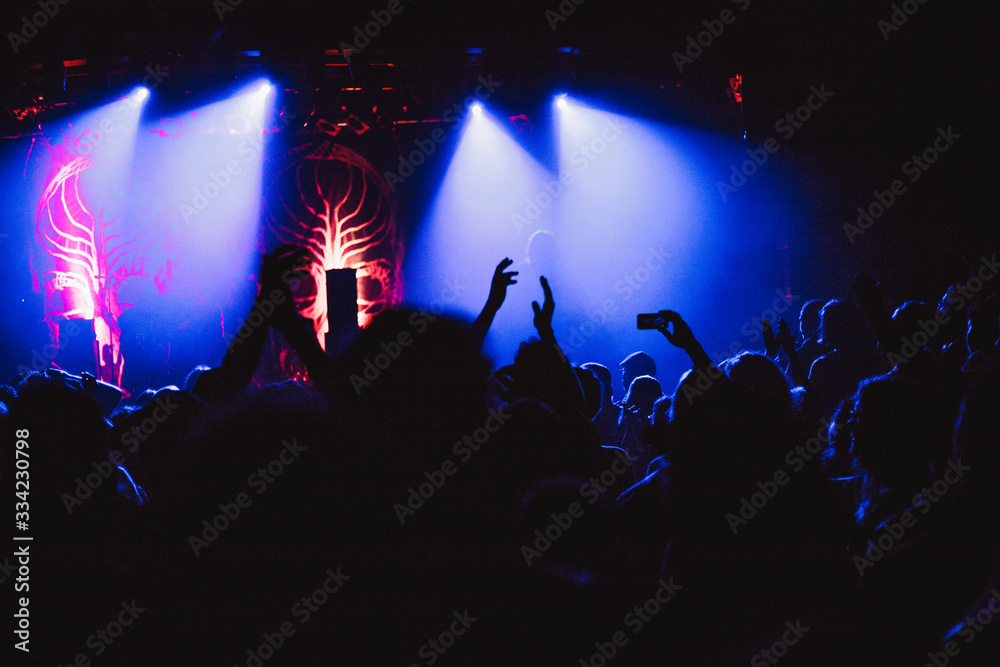Concert crowd attending a concert, people silhouettes are visible, backlit by stage lights, raised hands and smart phones are visible.