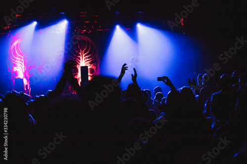 Concert crowd attending a concert, people silhouettes are visible, backlit by stage lights, raised hands and smart phones are visible.