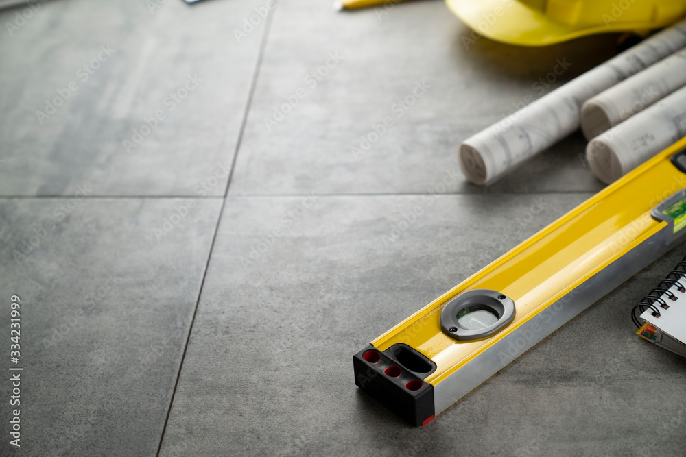Contractor concept. Tool kit of the contractor: yellow hardhat, libella, hand saw, and notebook on the gray tiles background. Contractor concept. Tool kit of the contractor: yellow hardhat, libella, hand saw, and notebook on the gray tiles background.