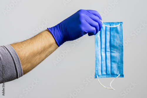 A hand in protective medical glove hold a disposable medical mask on a gray background