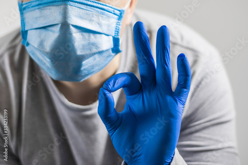 A hand in a protective medical glove shows an okay gesture near face in a protective mask