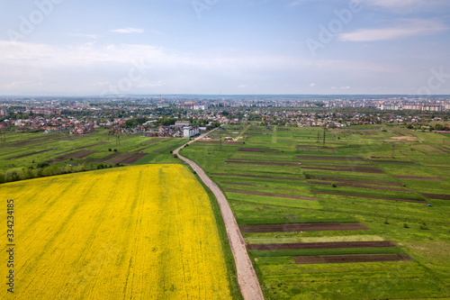 Aerial view of ground road with moving cars in green fields with blooming rapeseed plants  suburb houses on horizon and blue sky copy space background. Drone photography.