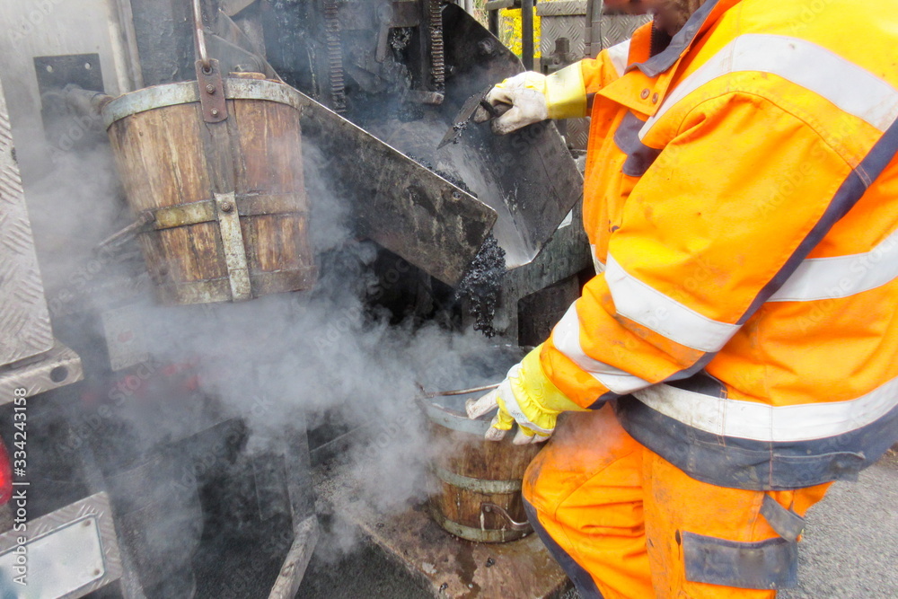Road construction, road rehabilitation, asphalting. A worker in protective suit fills boiling hot asphalt from an asphalt machine into a bucket to fill a pothole. Road construction, road rehabilitation, asphalting. A worker in protective suit fills boiling hot asphalt from an asphalt machine into a bucket to fill a pothole.