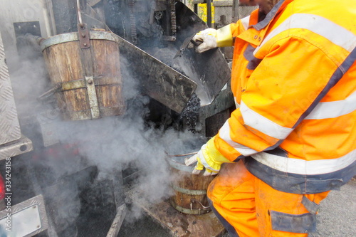 Road construction, road rehabilitation, asphalting. A worker in protective suit fills boiling hot asphalt from an asphalt machine into a bucket to fill a pothole.