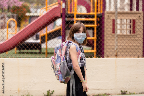 A school aged girl is longing to attend her closed school. She is in front of her school with a facemask on during the covid-19 outbreak.
