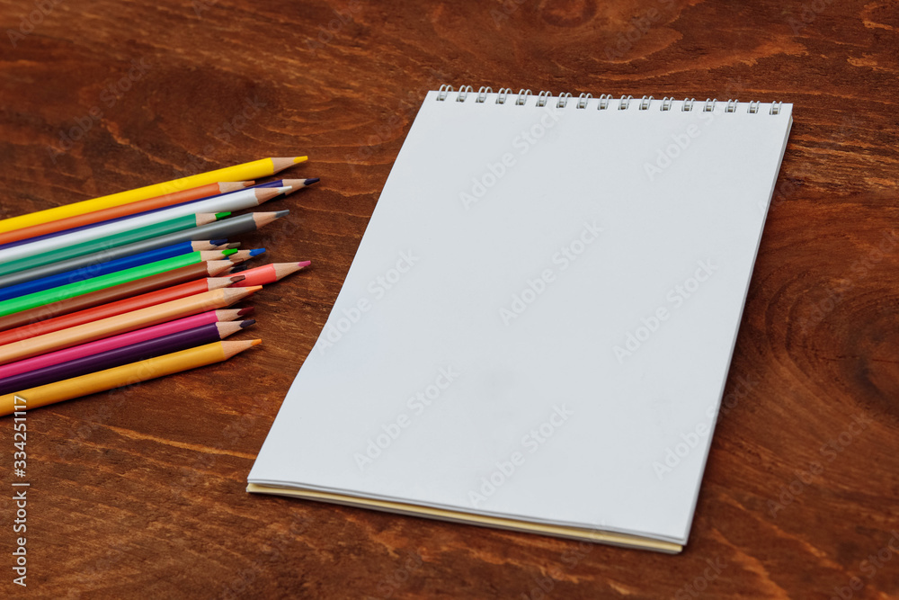 A Notepad with white sheets and an empty space for text on a spring and colored pencils next to it on a brown wooden table A Notepad with white sheets and an empty space for text on a spring and colored pencils next to it on a brown wooden table