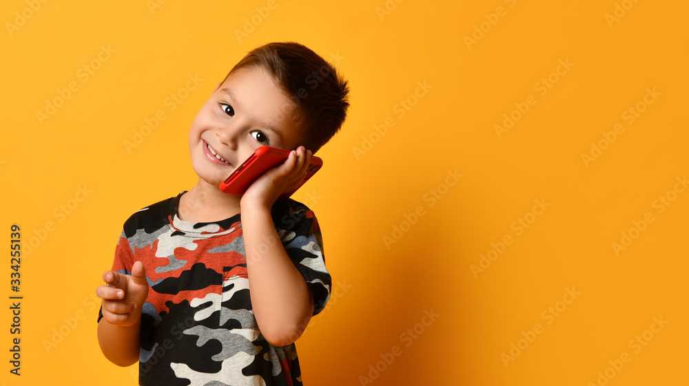 Little brunet boy in camouflage t-shirt is smiling, talking by red smartphone, looking at you, posing against orange background Little brunet boy in camouflage t-shirt is smiling, talking by red smartphone, looking at you, posing against orange background