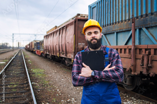 Portrait of railroader foreman holding checklist and controlling cargo dispatch. Freight train with shipping containers in background. Railway transportation.
