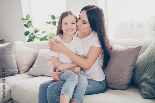 Photo of pretty little girl young charming mommy hugging holding each other close affectionate kissing daughter cheek sitting comfy sofa spend time together indoors home house