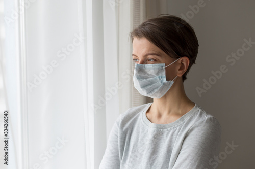 Portrait of a sad pensive young woman in medical mask in quarantine near the window.