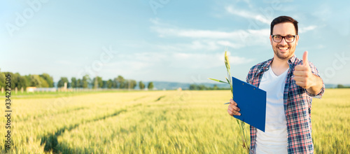 Happy young millennial Caucasian farmer or agronomist inspecting wheat fields and filling questionnaire, showing thumbs up and looking directly at camera