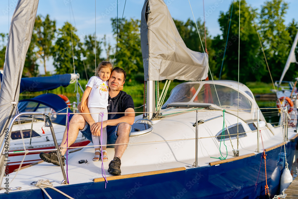 Portrait of smiling father and daughter on prow of sailboat or yacht anchored in marina at bright sunny day. Blurred background with boats and trees Portrait of smiling father and daughter on prow of sailboat or yacht anchored in marina at bright sunny day. Blurred background with boats and trees