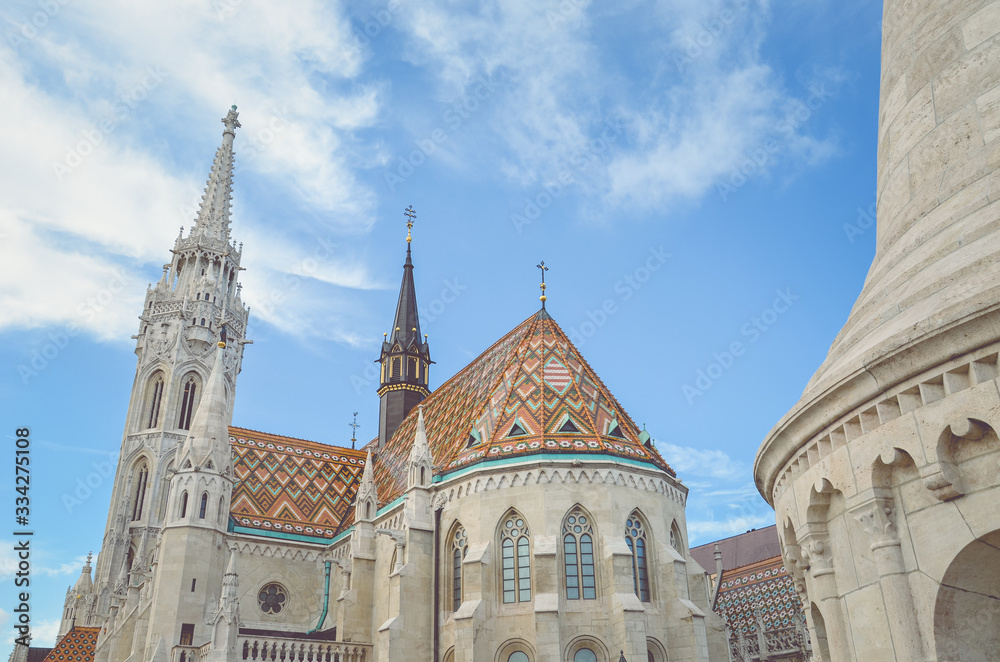 The spire of the famous Matthias Church in Budapest, Hungary. Roman Catholic church built in the Gothic style. Orange colored tile roof. Blue sky and white clouds above. Horizontal photo with filter