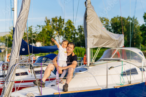 Portrait of smiling father and daughter on prow of sailboat or yacht anchored in marina at bright sunny day. Blurred background with boats and trees