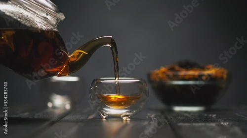 Tea ceremony. The woman's hand pours hot black tea with flowers from a transparent teapot into glass piala cups. Black background, counter light.