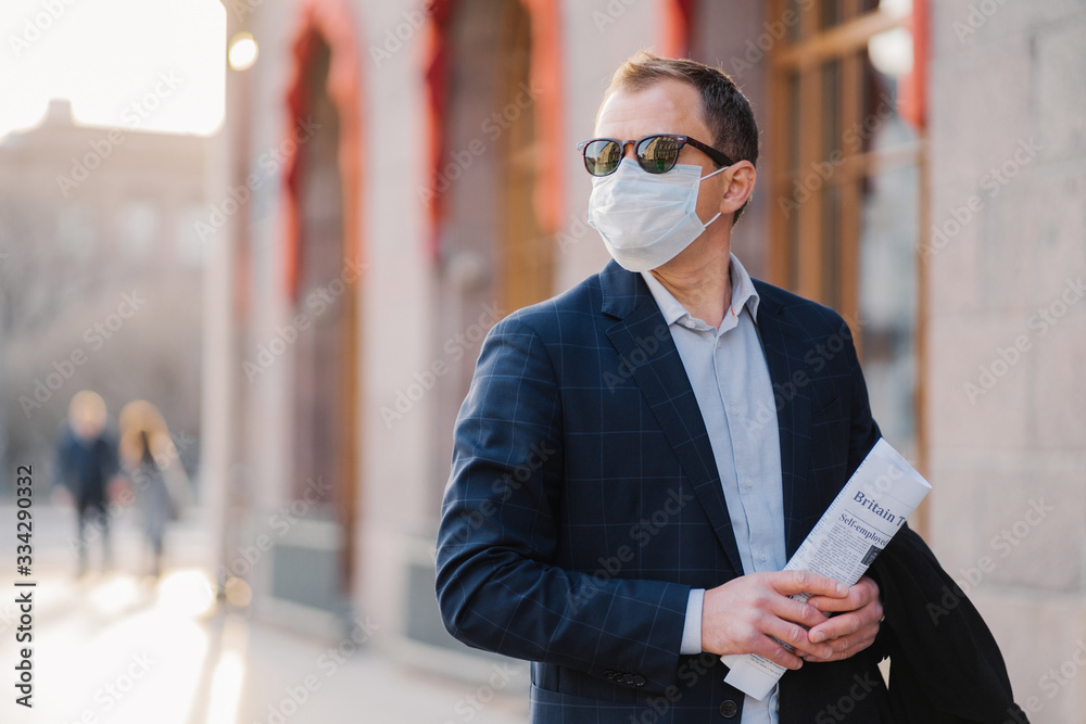 Working in pandemic situation. Business worker or entrepreneur wears medical mask for coronavirus protection stands beside office building outdoor holds newspapers concentrated somewhere into distance - obrazy, fototapety, plakaty 
