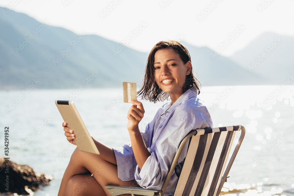 Smiling woman sitting on deck chair by the sea using tablet and credit card on a sunny day Smiling woman sitting on deck chair by the sea using tablet and credit card on a sunny day